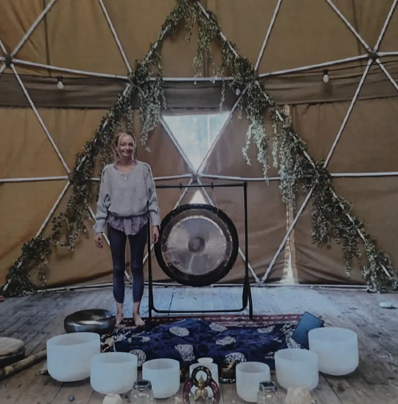 Patsy standing beside a gong inside a geodesic dome hung with greenery, crystal bowls laid out on the floor in front