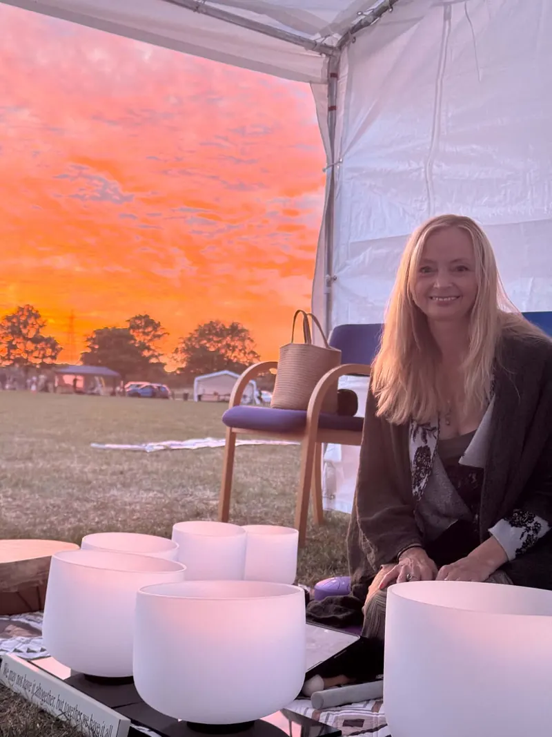 Patsy smiling beside a set of crystal singing bowls in a gazebo, with an orange sunset sky over a field behind her